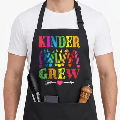 Crew 1st kindergarten teacher apron - KINDER GREW text with colorful pencils and arrow heart symbol in cartoon style — shown on a black cotton canvas apron against a white background
