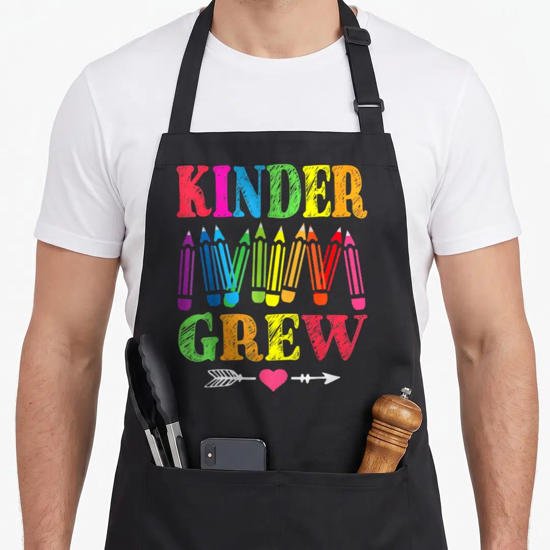 Crew 1st kindergarten teacher apron - KINDER GREW text with colorful pencils and arrow heart symbol in cartoon style — shown on a black cotton canvas apron against a white background