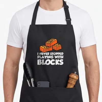 Masonry mason apron - Three orange bricks above white text I NEVER STOPPED PLAYING WITH BLOCKS — shown on a black cotton canvas apron against a white background