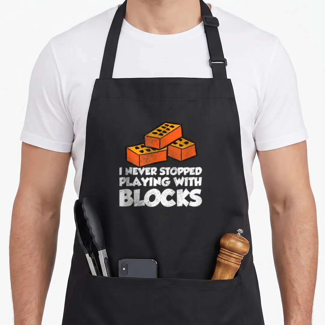 Masonry mason apron - Three orange bricks above white text I NEVER STOPPED PLAYING WITH BLOCKS — shown on a black cotton canvas apron against a white background