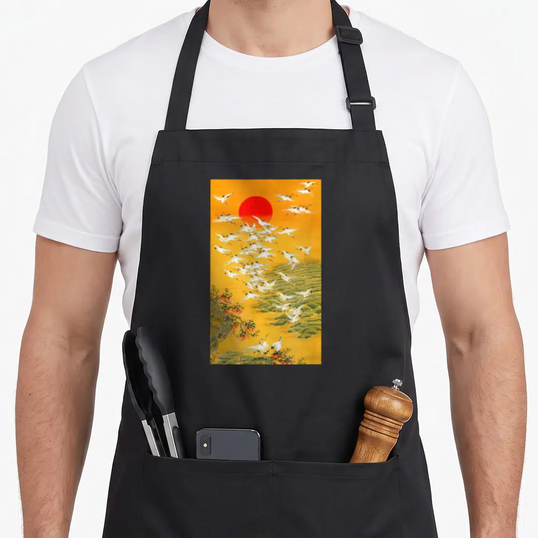 Japanese crane apron - Red sun over yellow sky with white cranes, green clouds, and fruit tree — shown on a black cotton canvas apron against a white background