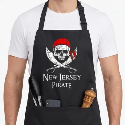 Jersey skull apron - White skull red bandana two cutlasses New Jersey Pirate distressed text — shown on a black cotton canvas apron against a white background