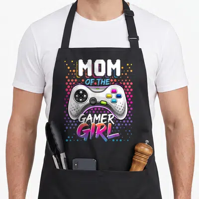 Gamer birthday video game apron - White game controller with MOM OF THE GAMER GIRL text over pink purple blue yellow — shown on a black cotton canvas apron against a white background