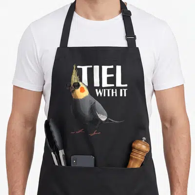 Cute parrot bird apron - Cockatiel wearing sunglasses next to stacked white text TIEL WITH IT — shown on a black cotton canvas apron against a white background