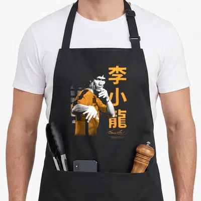 Fight iconic aprons - Stylized man in orange shirt next to vertical orange kanji and grey brick pattern — shown on a black cotton canvas apron against a white background