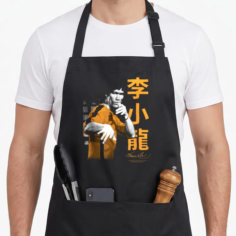 Fight iconic aprons - Stylized man in orange shirt next to vertical orange kanji and grey brick pattern — shown on a black cotton canvas apron against a white background