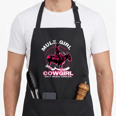 Vintage mule apron - Pink and black cartoon woman riding a mule with Mule Girl Just Like a Cowgirl text — shown on a black cotton canvas apron against a white background
