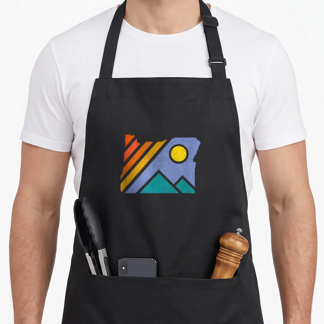 Throwback oregon dunes apron - Oregon state map silhouette with red orange yellow blue stripes and teal mountains — shown on a black cotton canvas apron against a white background