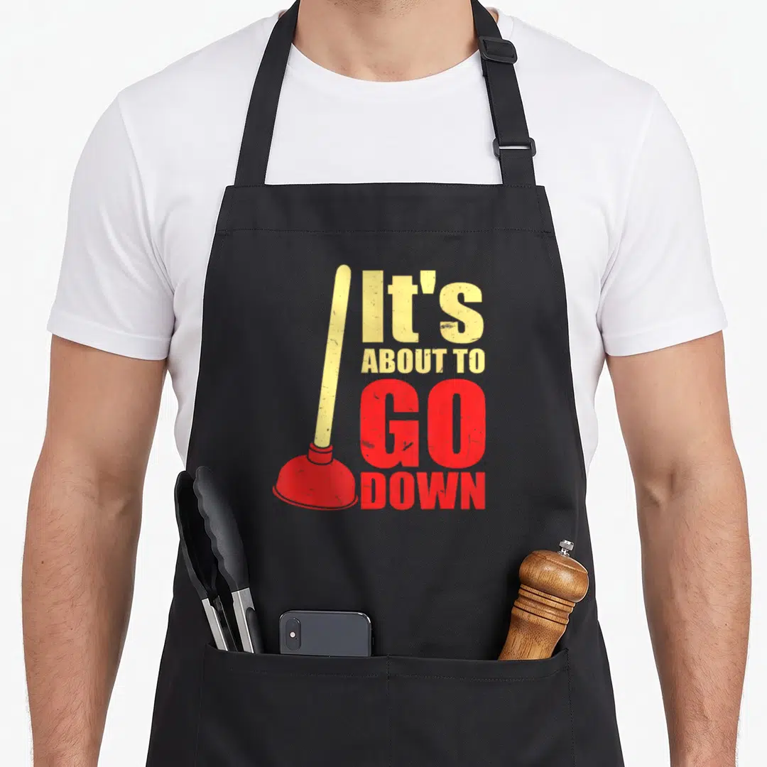 Plunger joke apron - Red plunger and yellow distressed text reading It's About To Go Down — shown on a black cotton canvas apron against a white background