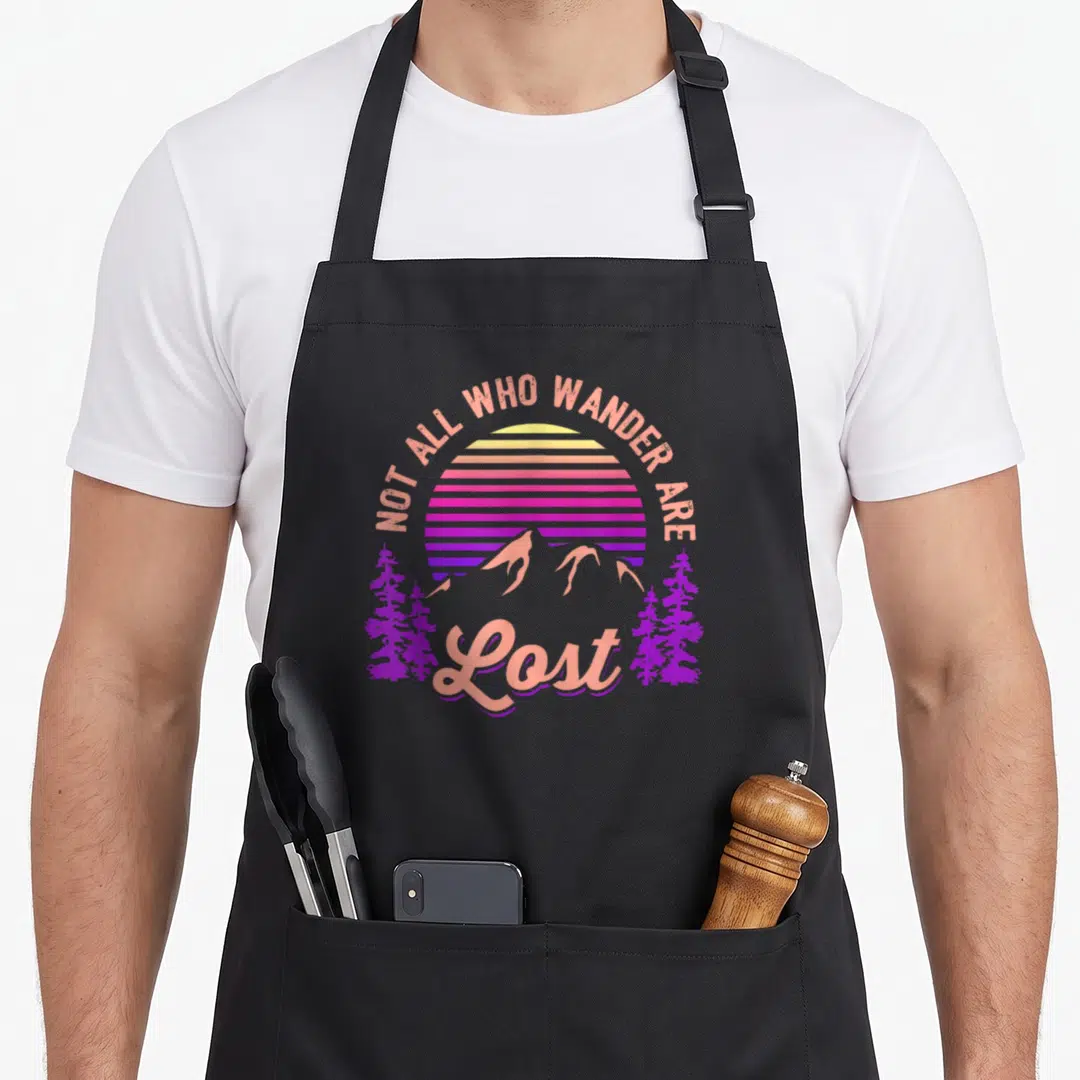 Wander mountain girl apron - Retro sunset mountains and trees with text NOT ALL WHO WANDER ARE Lost in purple pink — shown on a black cotton canvas apron against a white background