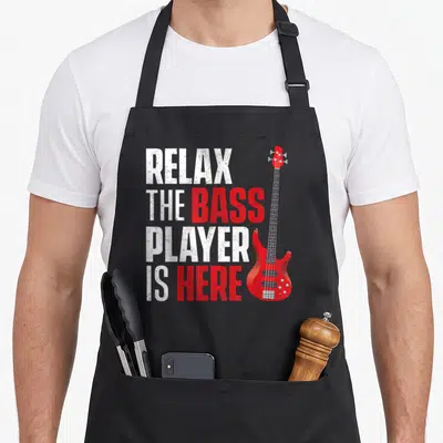 Musician bass apron - Red bass guitar next to stacked text RELAX THE BASS PLAYER IS HERE in distressed white and red — shown on a black cotton canvas apron against a white background