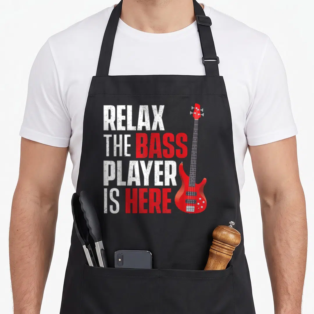 Musician bass apron - Red bass guitar next to stacked text RELAX THE BASS PLAYER IS HERE in distressed white and red — shown on a black cotton canvas apron against a white background