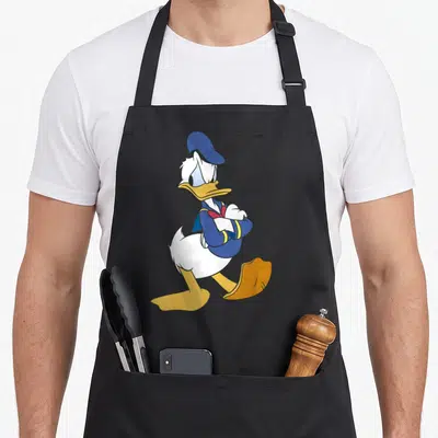 Traditional duck apron - Cartoon duck walking with arms crossed wearing blue hat and coat with yellow trim — shown on a black cotton canvas apron against a white background