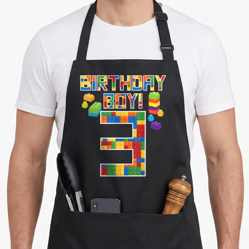 3rd birthday building aprons - Colorful brick pattern letters spelling BIRTHDAY BOY! above a large number 3 — shown on a black cotton canvas apron against a white background