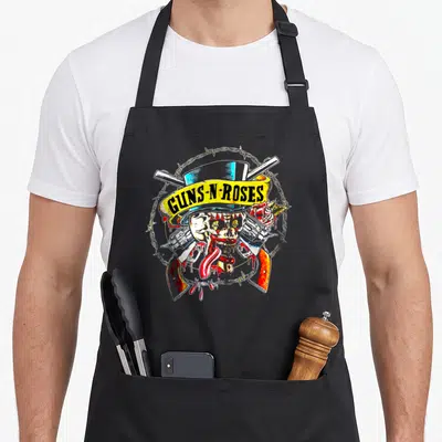 Cool rose gun apron - Skull with tongue, crossed guns, roses, barbed wire circle, yellow GUNS-N-ROSES banner — shown on a black cotton canvas apron against a white background