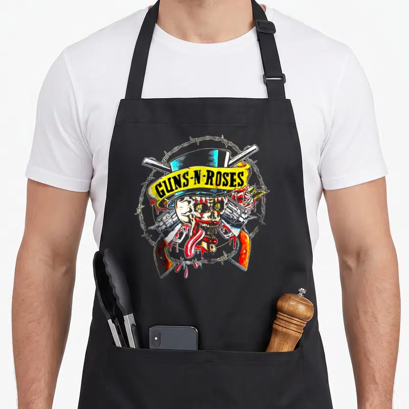 Cool rose gun apron - Skull with tongue, crossed guns, roses, barbed wire circle, yellow GUNS-N-ROSES banner — shown on a black cotton canvas apron against a white background