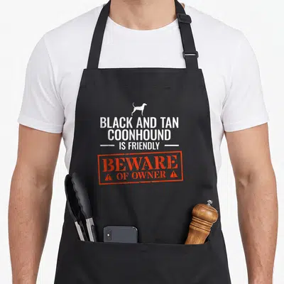 Coonhound black apron - White dog silhouette above text BEWARE OF OWNER in orange box — shown on a black cotton canvas apron against a white background