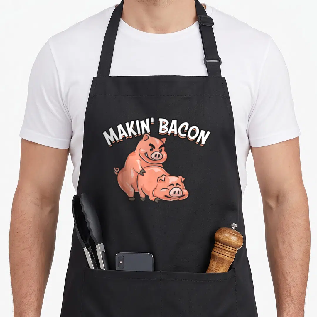 Bacon making an apron - Arched MAKIN BACON text above standing and lying pink cartoon pigs — shown on a black cotton canvas apron against a white background