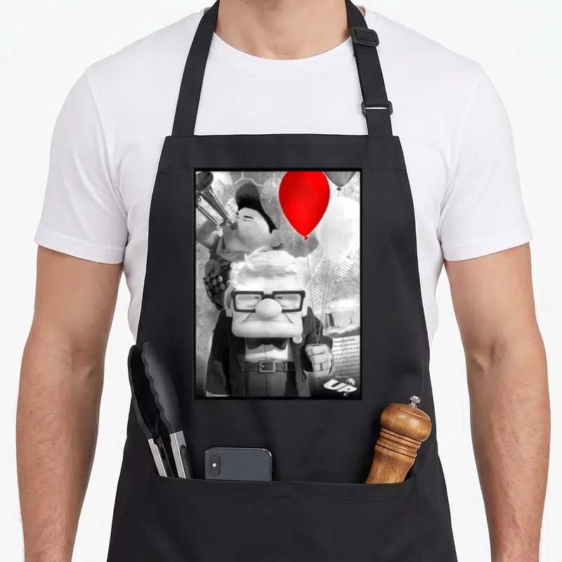 Carl trumpet apron - Carl Fredricksen holds red balloon while Russell plays trumpet behind him with UP logo — shown on a black cotton canvas apron against a white background