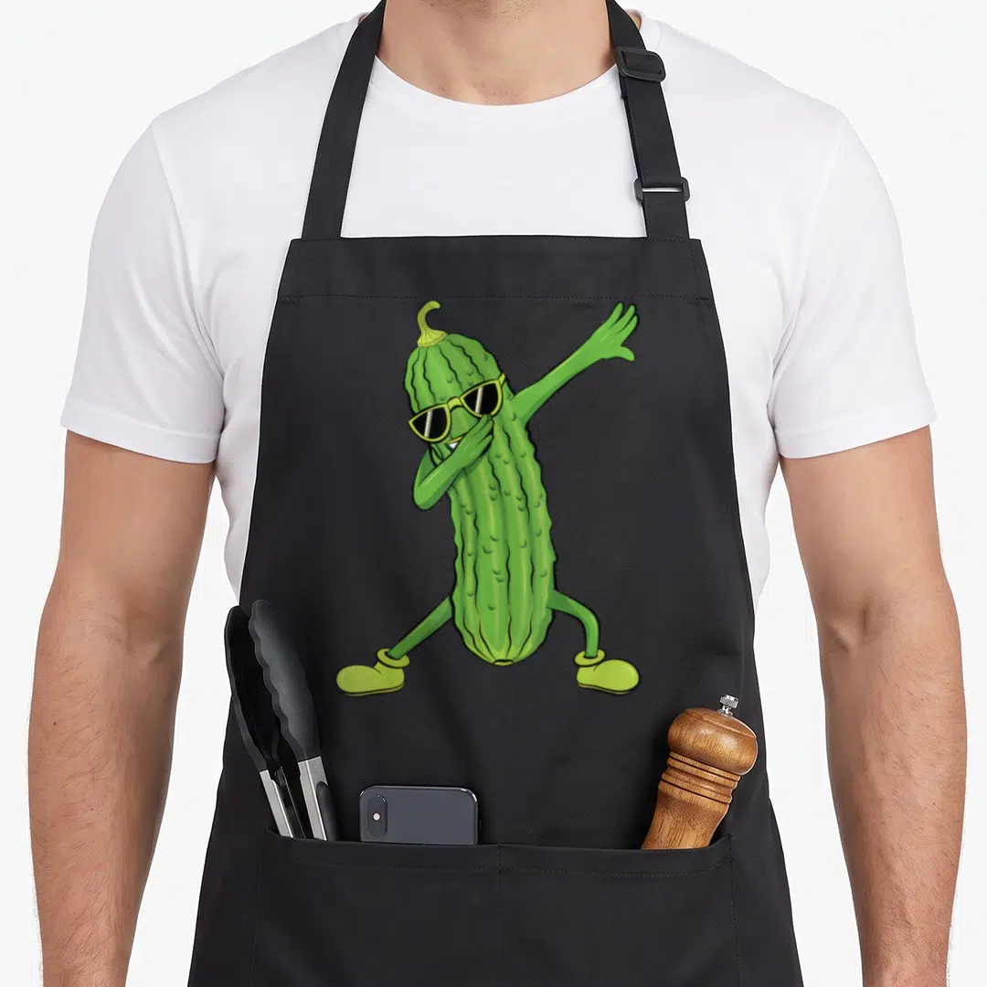 Dabbing pickle apron - Cartoon green pickle wearing sunglasses and shoes in a dab pose — shown on a black cotton canvas apron against a white background