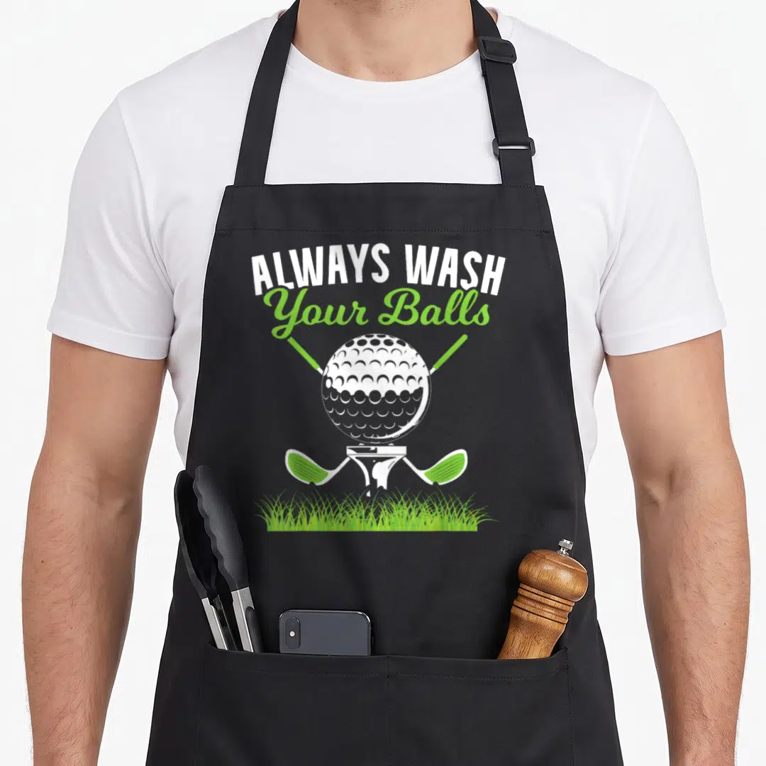 Wash golf apron - Arched ALWAYS WASH Your Balls text above golf ball on tee between crossed clubs with grass — shown on a black cotton canvas apron against a white background