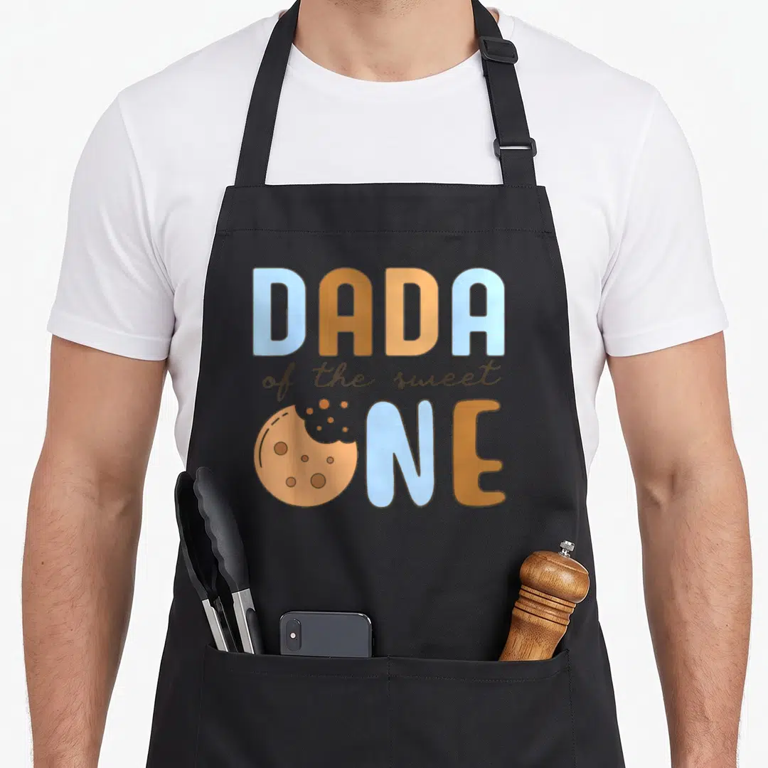 Dada cookies apron - DADA of the sweet ONE text with bitten cookie graphic in blue and tan — shown on a black cotton canvas apron against a white background