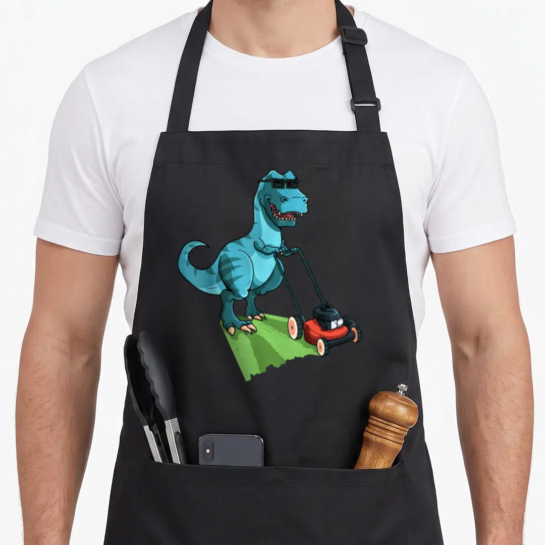 Mower t-rex apron - Blue cartoon dinosaur wearing sunglasses pushing red lawnmower on green grass patch — shown on a black cotton canvas apron against a white background