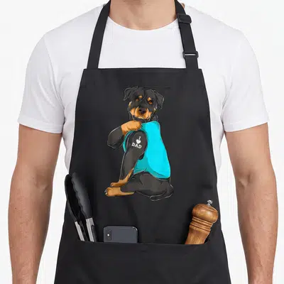 Rottweiler tattoo aprons - Cartoon rottweiler in cyan shirt showing I heart dad text on arm — shown on a black cotton canvas apron against a white background