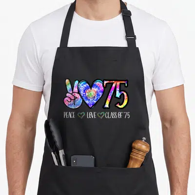50th tie-dye apron - Tie-dye peace sign, heart, and 75 with PEACE LOVE CLASS OF '75 text — shown on a black cotton canvas apron against a white background