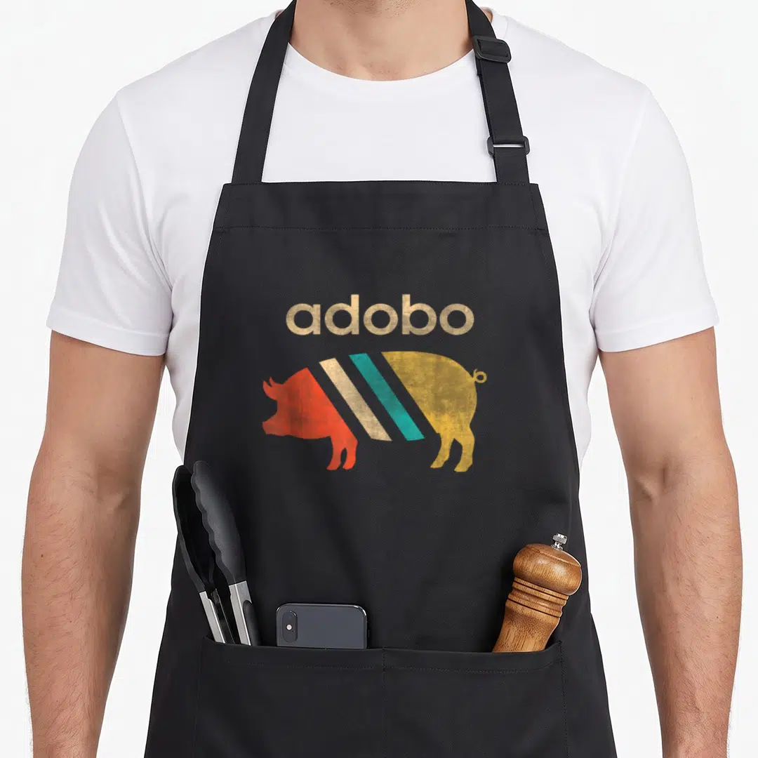 Pork filipino apron - adobo text above a pig silhouette filled with tan red teal and yellow diagonal stripes — shown on a black cotton canvas apron against a white background