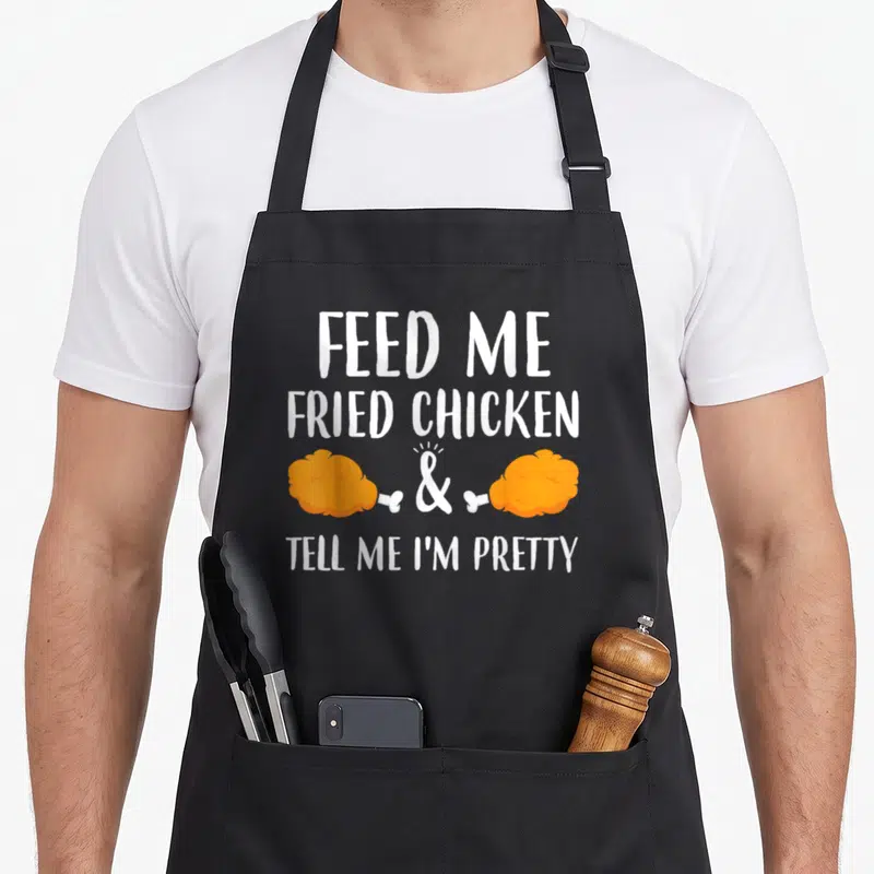Feed chicken apron - White text FEED ME FRIED CHICKEN & TELL ME I'M PRETTY with two orange drumsticks — shown on a black cotton canvas apron against a white background