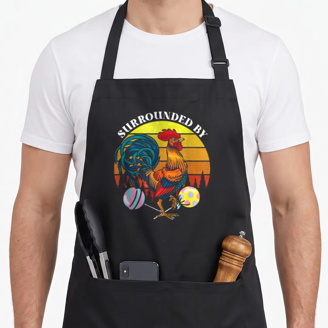 Surrounded rooster apron - Cartoon rooster balancing two patterned balls under arched text SURROUNDED BY against — shown on a black cotton canvas apron against a white background