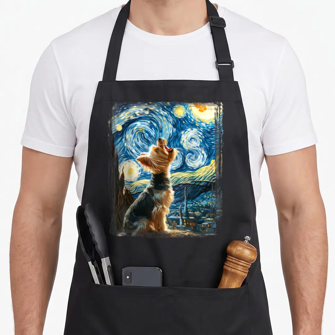 Artistic yorkie apron - Howling Yorkshire Terrier set against swirling blue night sky and village lights — shown on a black cotton canvas apron against a white background