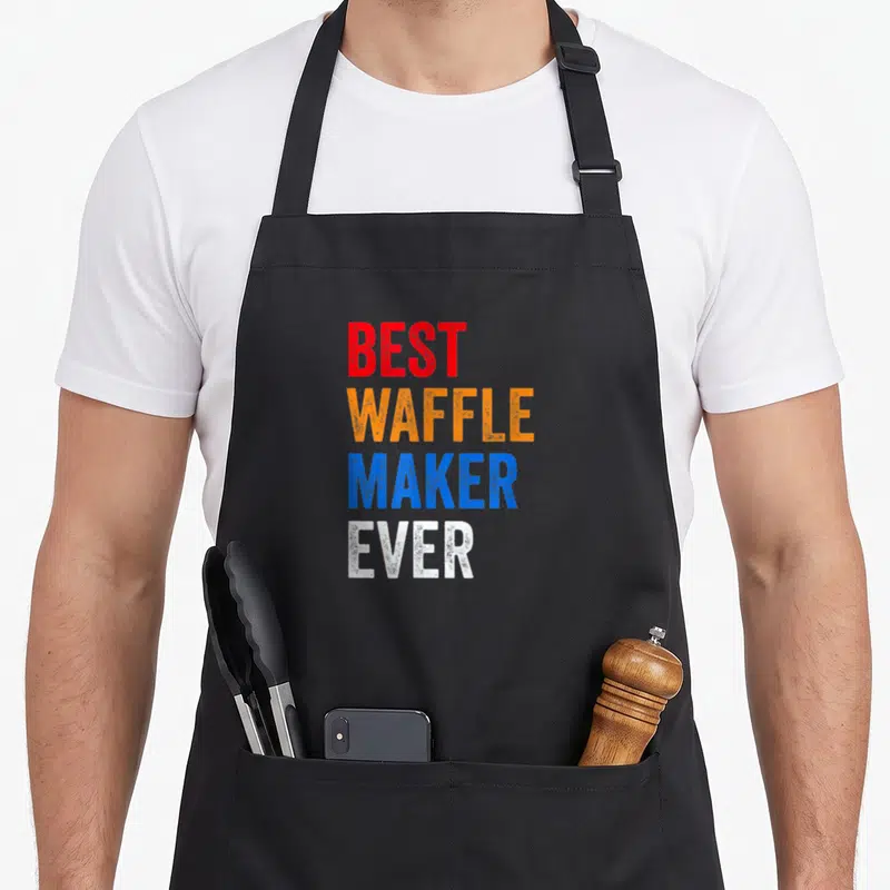 Waffle baker makers apron - Stacked text BEST WAFFLE MAKER EVER in red orange blue and white block letters — shown on a black cotton canvas apron against a white background
