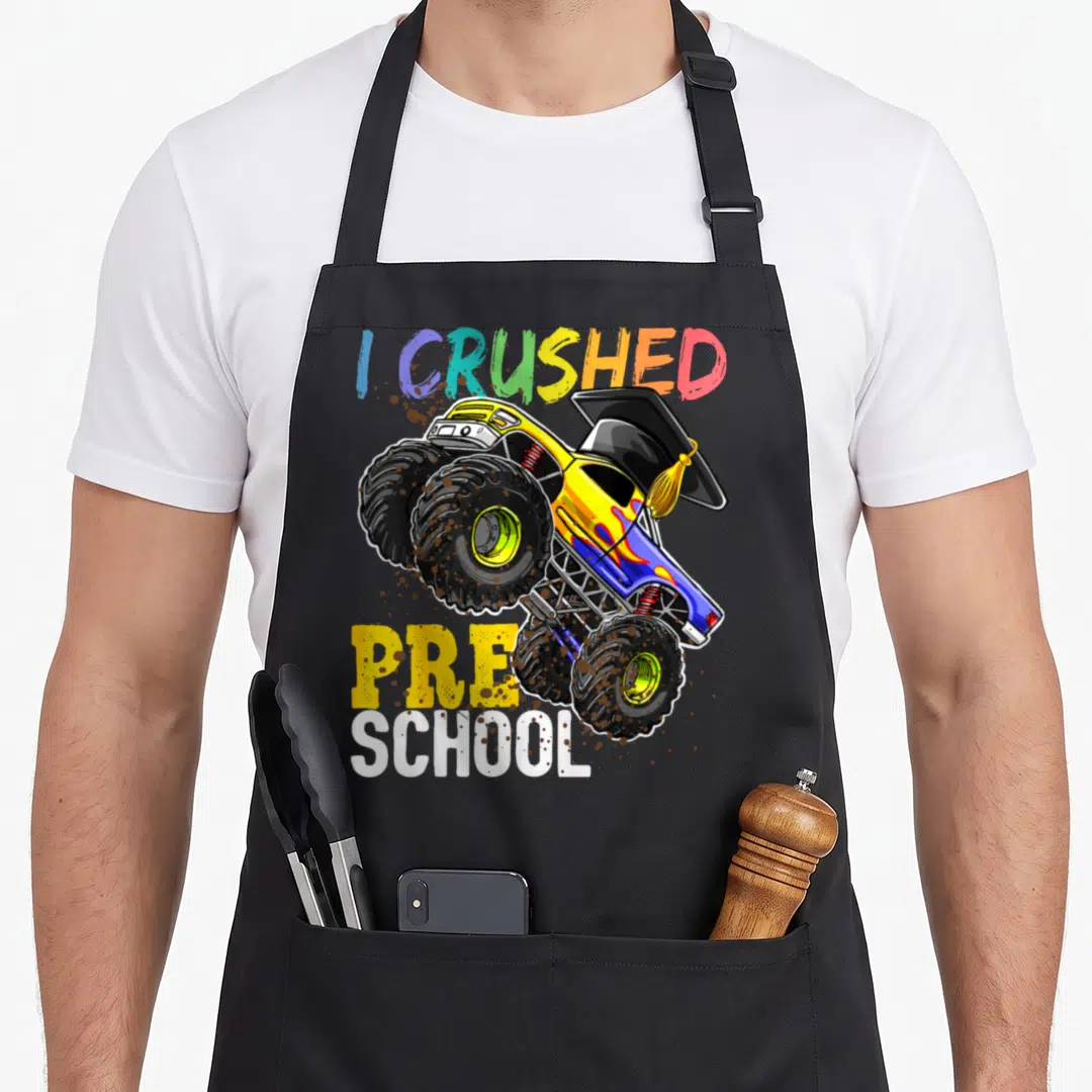 Crushed truck apron - Cartoon monster truck with graduation cap and I Crushed Pre School text — shown on a black cotton canvas apron against a white background