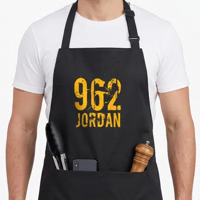 Funny area code apron - Orange distressed text reading 962 JORDAN stacked vertically — shown on a black cotton canvas apron against a white background