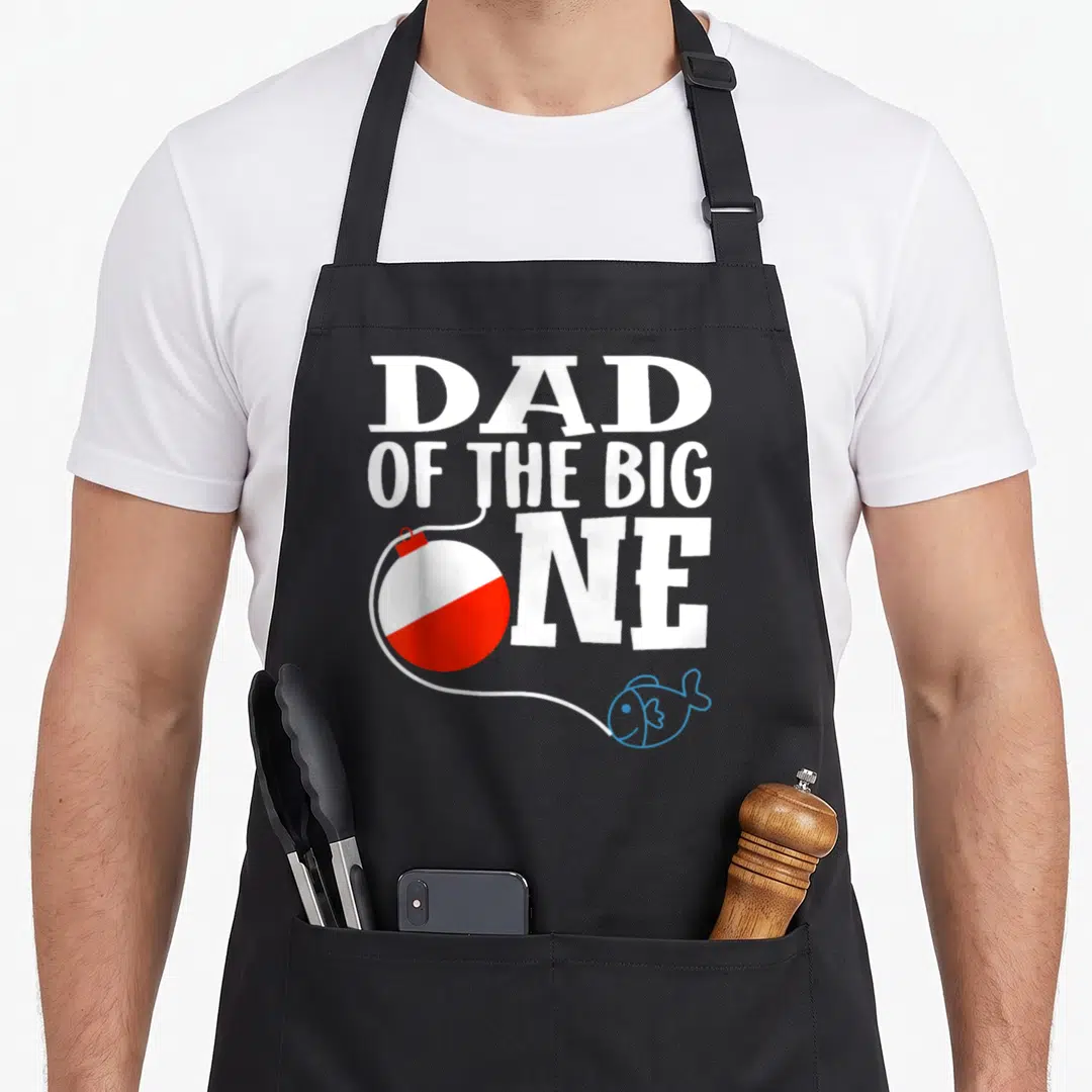 Birthday fishing aprons - Dad of the Big One text with red white bobber and blue fish — shown on a black cotton canvas apron against a white background