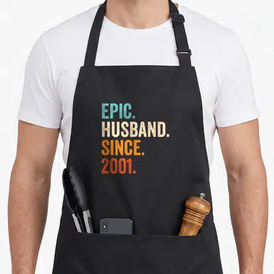 25th wedding apron - Stacked EPIC HUSBAND SINCE 2001 text in teal beige and orange distressed typography — shown on a black cotton canvas apron against a white background