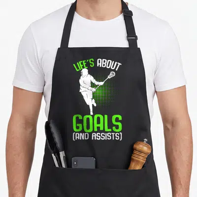 Youth lacrosse player apron - White lacrosse player running with stick between LIFE'S ABOUT GOALS text and green — shown on a black cotton canvas apron against a white background