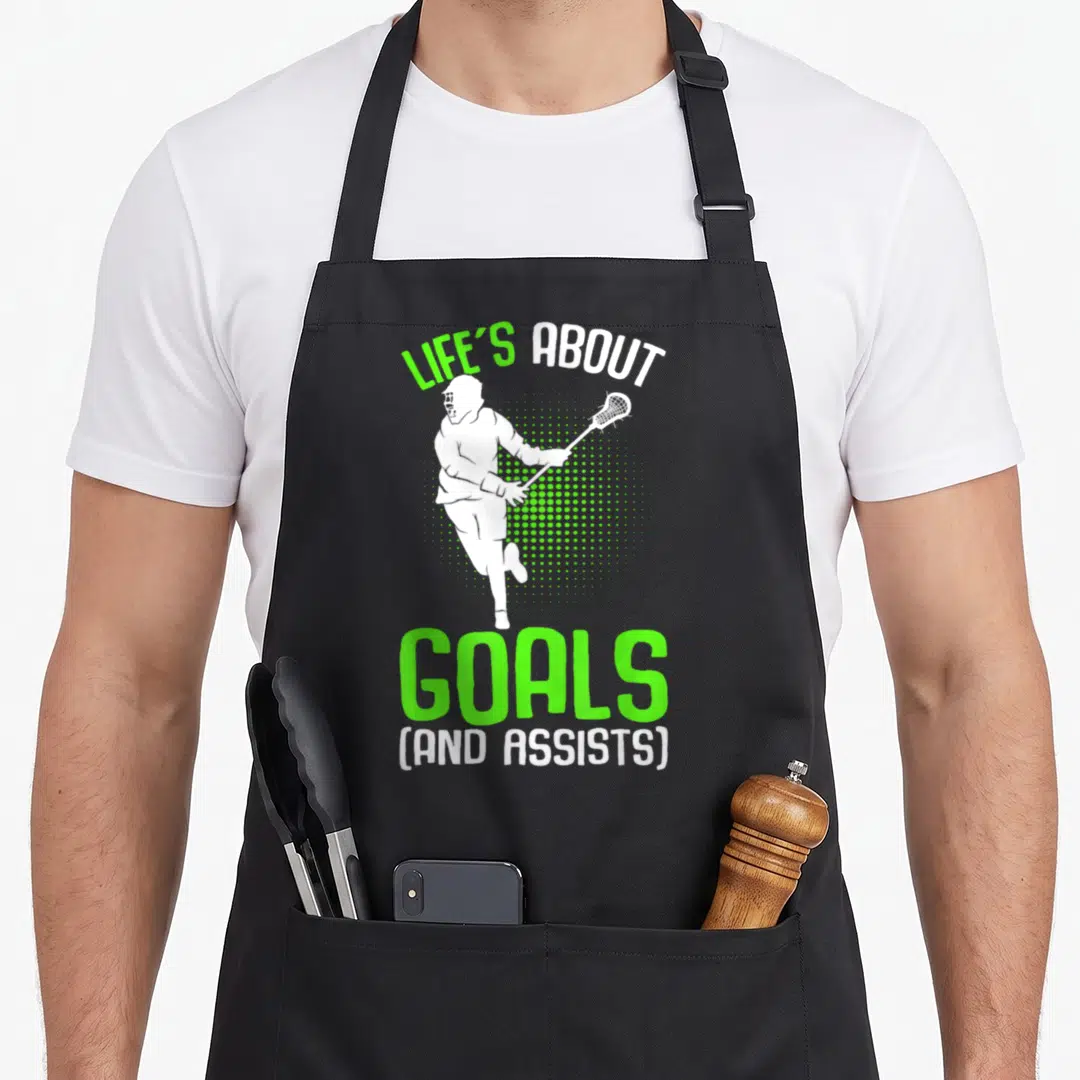Youth lacrosse player apron - White lacrosse player running with stick between LIFE'S ABOUT GOALS text and green — shown on a black cotton canvas apron against a white background
