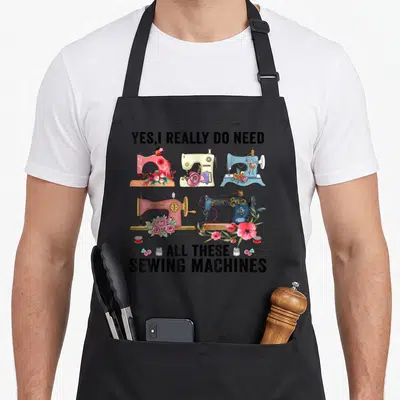 Floral do i need an apron - Five cartoon sewing machines in pink blue and cream with flowers and bold black text — shown on a black cotton canvas apron against a white background