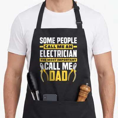 Father lineman electricians apron - Stacked text reading Call Me Dad flanked by two yellow and grey pliers on grey — shown on a black cotton canvas apron against a white background