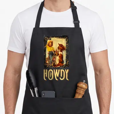 Rustic southern aprons - Retro cowgirl and horse illustration in leopard frame above HOWDY text — shown on a black cotton canvas apron against a white background