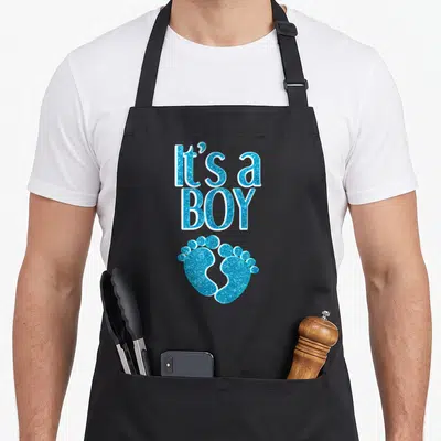 Pregnancy pregnant apron - Stacked It's a BOY text above two blue baby footprints with white outlines — shown on a black cotton canvas apron against a white background