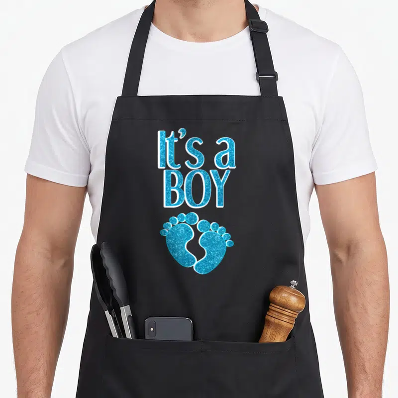 Pregnancy pregnant apron - Stacked It's a BOY text above two blue baby footprints with white outlines — shown on a black cotton canvas apron against a white background