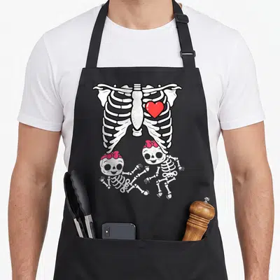 Skeleton x ray apron - Cartoon ribcage with red heart above two baby skeletons wearing red bows — shown on a black cotton canvas apron against a white background