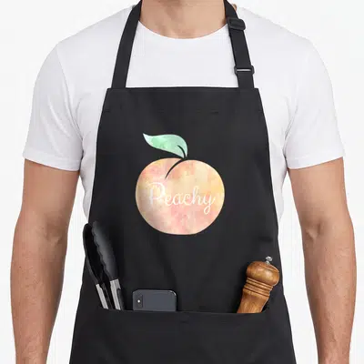 Peachy peaches apron - Watercolor peach in orange and pink with green leaf and white Peachy script text — shown on a black cotton canvas apron against a white background