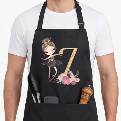 7th ballerina apron - Cartoon ballerina in black tutu next to glittery gold number 7 with pink and purple flowers — shown on a black cotton canvas apron against a white background