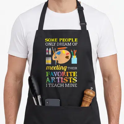 Dream teacher apron - Art supplies cluster with text Some People Only Dream Of Meeting Their Favorite Artists I Teach — shown on a black cotton canvas apron against a white background