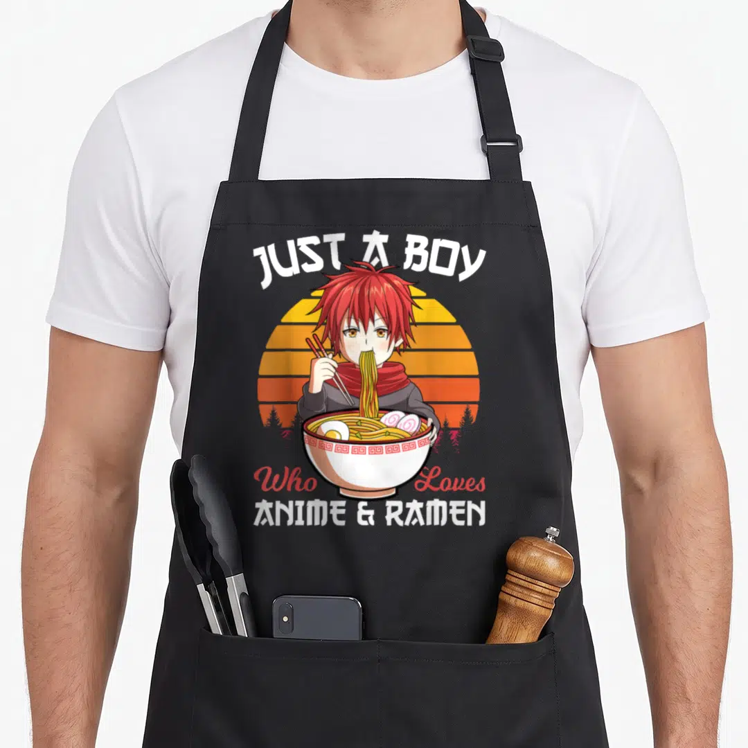 Ramen japanese anime apron - Red-haired cartoon boy eating ramen bowl under retro striped sun with white and red text — shown on a black cotton canvas apron against a white background
